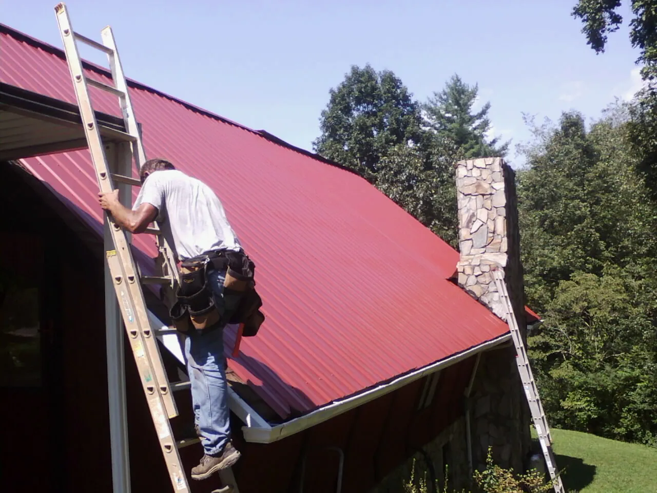 Roofer on a ladder beside a red metal roof and stone chimney — Soffit & Fascia Repair by Roofing Brothers in Kings Mills